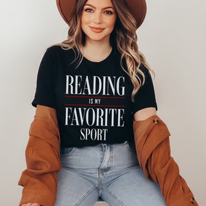 Może przedstawiać: Czarny T-shirt z białym napisem "Reading is my favorite sport". Koszulkę nosi osoba z długimi brązowymi włosami i brązowym kapeluszem.