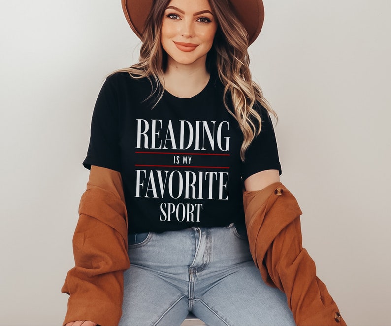 Może przedstawiać: Czarny T-shirt z białym napisem "Reading is my favorite sport". Koszulkę nosi osoba z długimi brązowymi włosami i brązowym kapeluszem.