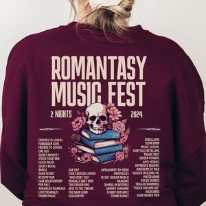 May include: A burgundy sweatshirt featuring "ROMANTASY MUSIC FEST 2024" text. The design includes a skull and flowers over books. The back lists romance novel themes and tropes, with "2 NIGHTS" displayed.