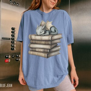 May include: Blue jean colored t-shirt featuring a sleeping baby dragon on a stack of books. The dragon is gray with white wings. The books are tan with gold-colored accents. The text "BLUE JEAN" is at the bottom left.