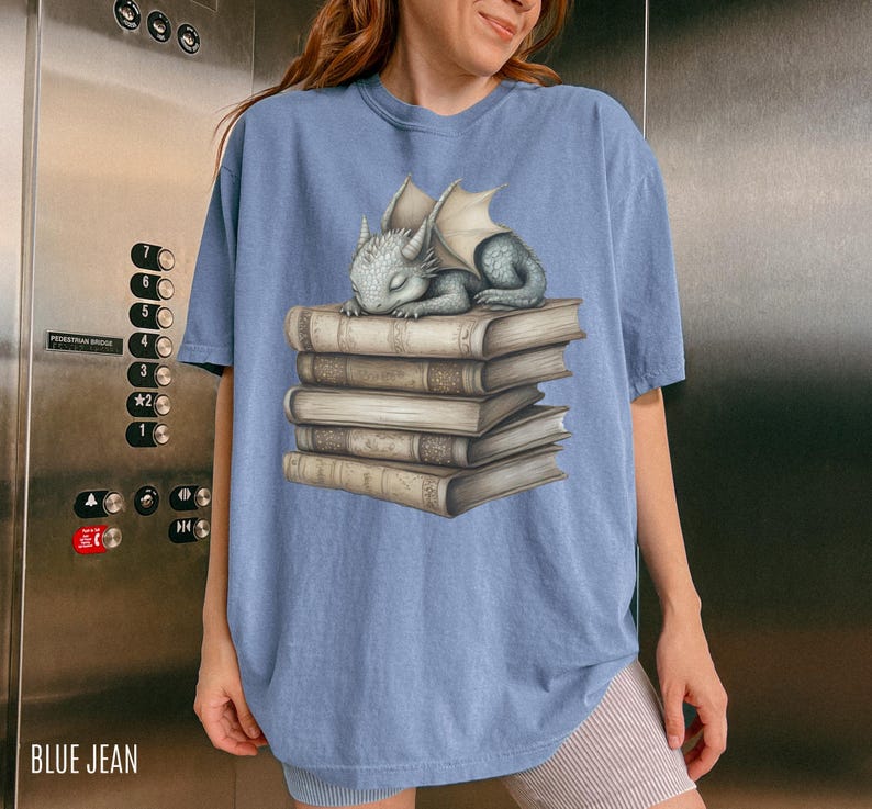 May include: Blue jean colored t-shirt featuring a sleeping baby dragon on a stack of books. The dragon is gray with white wings. The books are tan with gold-colored accents. The text "BLUE JEAN" is at the bottom left.