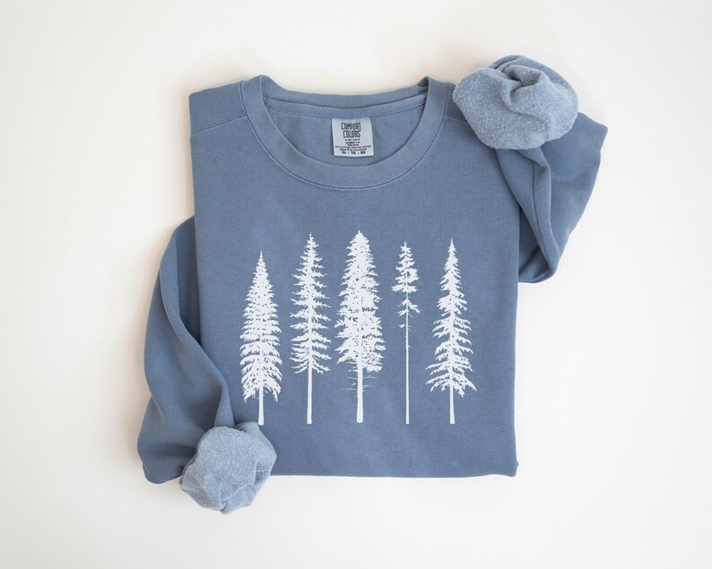 May include: A blue sweatshirt with a white print of five pine trees. The sweatshirt is folded and the sleeves are rolled up.