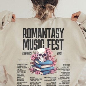 May include: Beige sweatshirt with the text "ROMANTASY MUSIC FEST 2024" and a skull, flowers, and books graphic. The design includes a list of romance novel tropes. A unique and stylish apparel item.