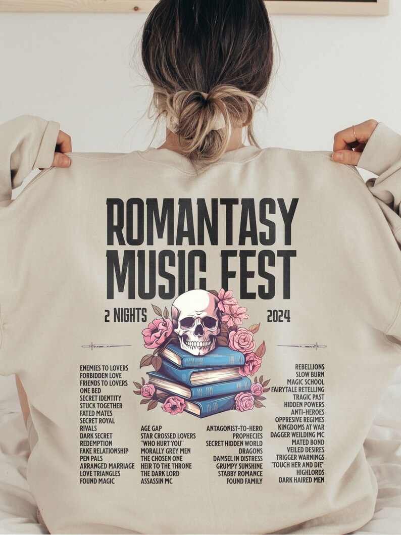 May include: Beige sweatshirt with the text "ROMANTASY MUSIC FEST 2024" and a skull, flowers, and books graphic. The design includes a list of romance novel tropes. A unique and stylish apparel item.