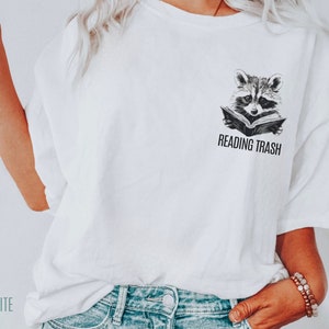 May include: White t-shirt featuring a black and white illustration of a raccoon reading a book. The text "READING TRASH" is printed below the raccoon.