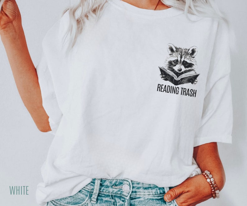 May include: White t-shirt featuring a black and white illustration of a raccoon reading a book. The text "READING TRASH" is printed below the raccoon.