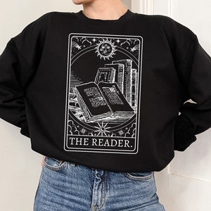 Puede incluir: Sudadera negra con un gr&aacute;fico blanco de una carta de tarot que representa un libro con el texto "THE READER."