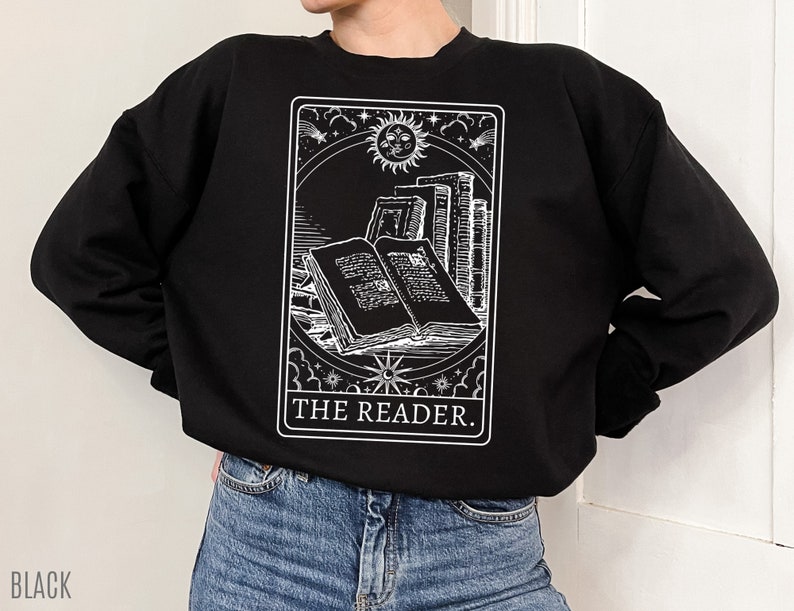 Puede incluir: Sudadera negra con un gr&aacute;fico blanco de una carta de tarot que representa un libro con el texto "THE READER."