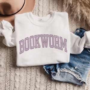 May include: A white sweatshirt with the word "BOOKWORM" in a purple collegiate font. The sweatshirt is folded on top of denim shorts. A beige hat and decorative plant are also in the image.