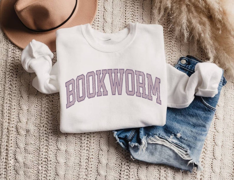 May include: A white sweatshirt with the word "BOOKWORM" in a purple collegiate font. The sweatshirt is folded on top of denim shorts. A beige hat and decorative plant are also in the image.