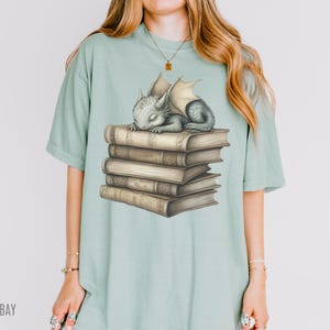 May include: A light green t-shirt featuring a sleeping baby dragon atop a stack of old books. The dragon is gray with cream-colored wings. The books are brown with gold-colored details. The word "BAY" is printed on the bottom left of the shirt.