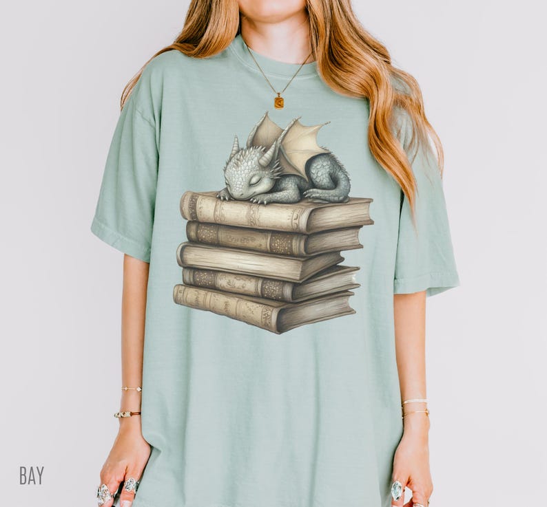 May include: A light green t-shirt featuring a sleeping baby dragon atop a stack of old books. The dragon is gray with cream-colored wings. The books are brown with gold-colored details. The word "BAY" is printed on the bottom left of the shirt.