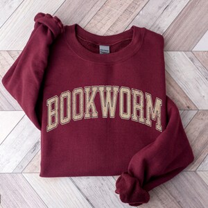 May include: Maroon sweatshirt with the word "BOOKWORM" in a beige, collegiate-style font. The sweatshirt is folded on a light wood-grain surface. The word "MAROON" is printed at the bottom left corner of the image.