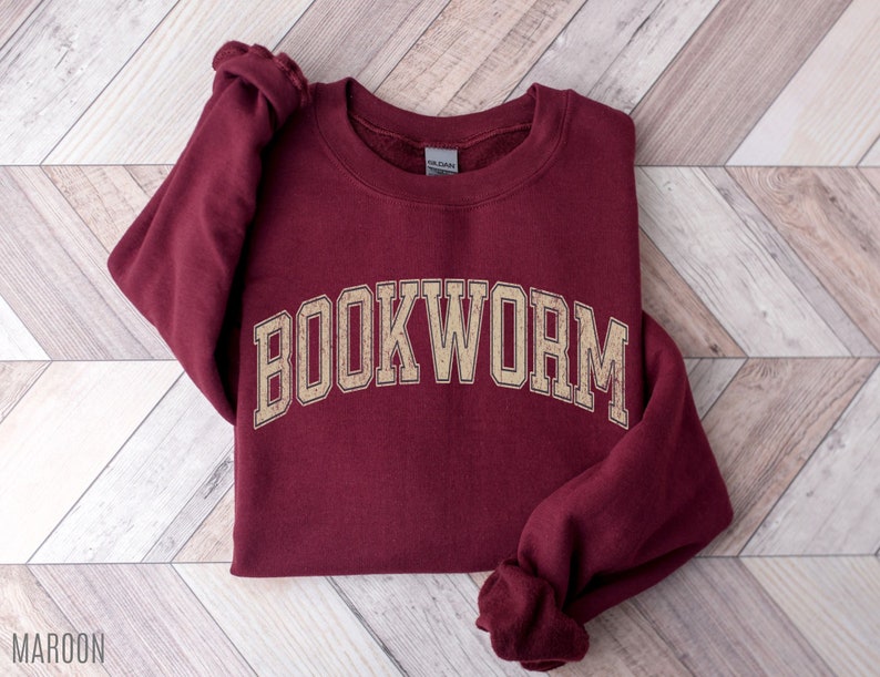 May include: Maroon sweatshirt with the word "BOOKWORM" in a beige, collegiate-style font. The sweatshirt is folded on a light wood-grain surface. The word "MAROON" is printed at the bottom left corner of the image.