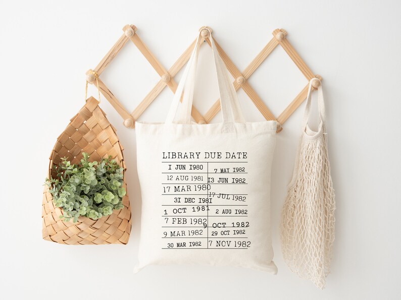 May include: A white canvas tote bag with black text that reads "LIBRARY DUE DATE" and a list of dates from 1980 to 1982. The bag is hanging on a wooden wall rack.