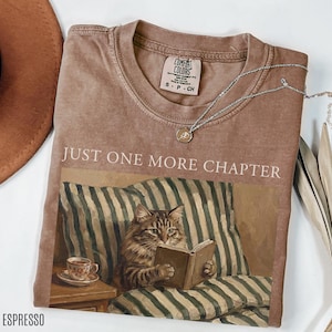 May include: Espresso-coloured t-shirt featuring the text "JUST ONE MORE CHAPTER" above an illustration of a cat reading a book. The cat is on a striped pillow. A silver necklace and a brown hat are also visible.