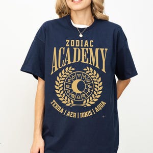 May include: Navy blue t-shirt with a gold graphic design. The design features the words "ZODIAC ACADEMY" above a circular emblem with zodiac symbols, a crescent moon, and the words "TERRA | AER | IGNIS | AQUA".