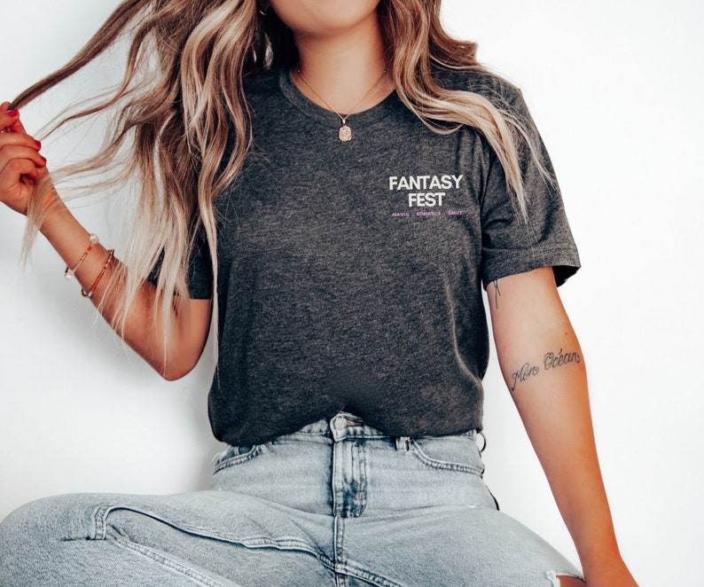 May include: A dark grey t-shirt with the text "Fantasy Fest" printed on the front. The text is in white with a purple outline. The shirt is being worn by a person with long blonde hair.