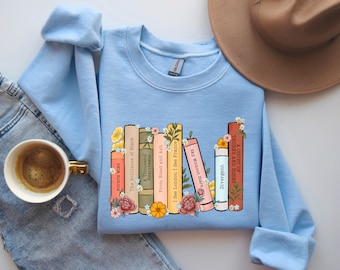 May include: Light blue sweatshirt with a graphic of a stack of books with floral accents. The book titles include "A Court of Thorns and Roses", "Divergent", "Sky Without Stars", "From Blood and Ash", "A Thousand Heartbeats", "The Brilliance of Stars", "Fourth Wing", and "I See London I See France".