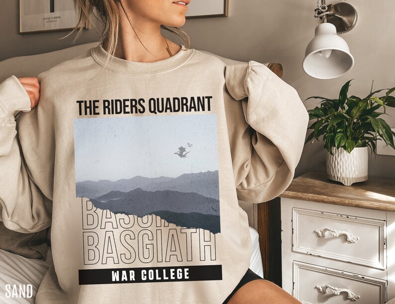 Puede incluir: Una sudadera beige con un estampado gr&aacute;fico de una cordillera y el texto "The Riders Quadrant Basgiath War College".
