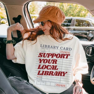 Woman wearing oversized cream t shirt with library card support your local library design sitting inside a car with hat and sunglasses