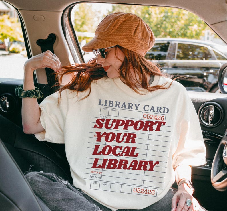 Woman wearing oversized cream t shirt with library card support your local library design sitting inside a car with hat and sunglasses