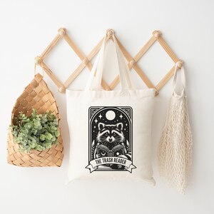 May include: Off-white tote bag featuring a black and white illustration of a raccoon reading a book under a night sky with stars and a moon. The text "THE TRASH READER" is on a banner below the raccoon.