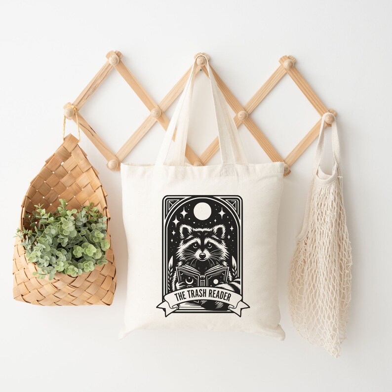 May include: Off-white tote bag featuring a black and white illustration of a raccoon reading a book under a night sky with stars and a moon. The text "THE TRASH READER" is on a banner below the raccoon.
