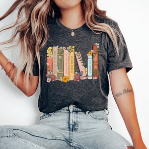 May include: A dark gray t-shirt with a graphic of a stack of books with floral accents. The book titles include "The Fault in Our Stars", "ACOTAR", "From Blood and Ash", "It Ends With Us", "Crescent City", and "A Court of Thorns and Roses".