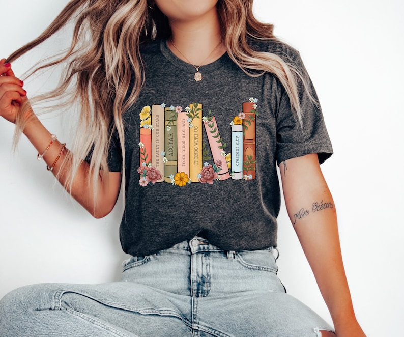 May include: A dark gray t-shirt with a graphic of a stack of books with floral accents. The book titles include "The Fault in Our Stars", "ACOTAR", "From Blood and Ash", "It Ends With Us", "Crescent City", and "A Court of Thorns and Roses".
