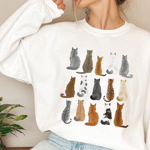 May include: White sweatshirt featuring a graphic print of numerous cats sitting in a grid pattern. The cats are of various colours and breeds.