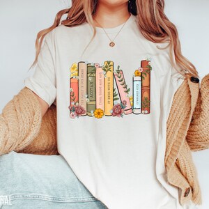 May include: A white t-shirt with a graphic of a stack of books with floral accents. The book titles are "The Fault in Our Stars", "ACOTAR", "From blood and ash", "It Ends With Us", "Crescent City".