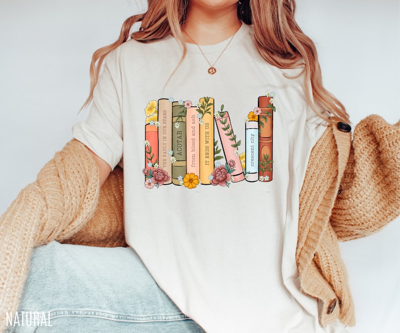May include: A white t-shirt with a graphic of a stack of books with floral accents. The book titles are "The Fault in Our Stars", "ACOTAR", "From blood and ash", "It Ends With Us", "Crescent City".