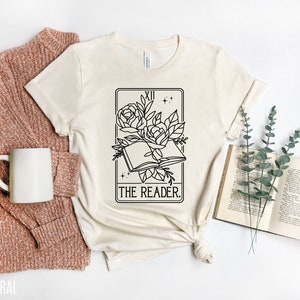 May include: Cream-colored t-shirt featuring a black and white tarot card design with a floral open book and the text "THE READER." The shirt is styled with a rust-colored sweater, a white mug, and an open book.