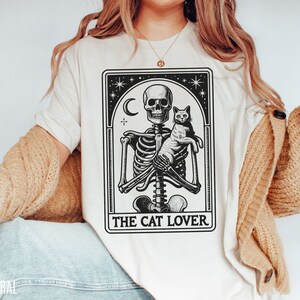 May include: Cream-colored t-shirt featuring a black and white graphic of a skeleton holding a cat, with a crescent moon and stars. The text "THE CAT LOVER" is printed below the image. The shirt has a relaxed fit.
