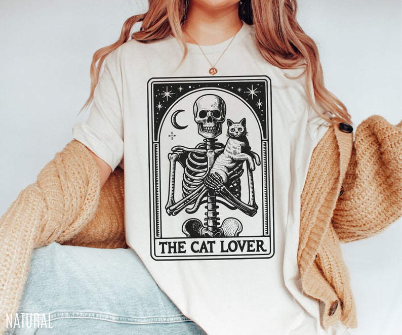 May include: Cream-colored t-shirt featuring a black and white graphic of a skeleton holding a cat, with a crescent moon and stars. The text "THE CAT LOVER" is printed below the image. The shirt has a relaxed fit.