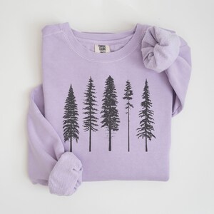 May include: A lavender sweatshirt with a black silhouette of a forest of five trees printed on the front. The sweatshirt has a crew neck and long sleeves. The text "Camping Club" is printed on a white tag sewn into the front of the sweatshirt.