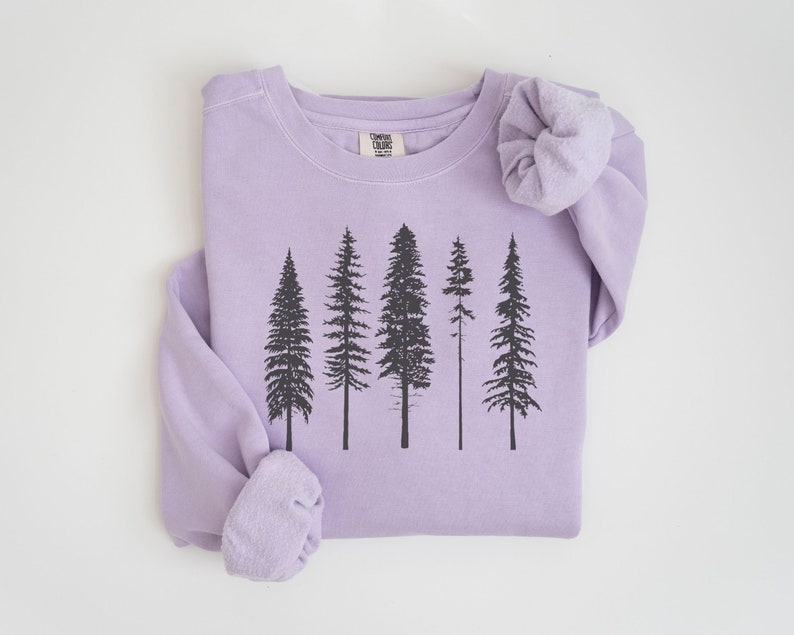 May include: A lavender sweatshirt with a black silhouette of a forest of five trees printed on the front. The sweatshirt has a crew neck and long sleeves. The text "Camping Club" is printed on a white tag sewn into the front of the sweatshirt.