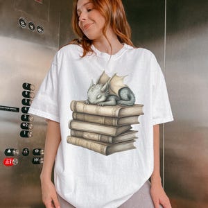 May include: White t-shirt featuring a sleeping baby dragon on a stack of books. The dragon is light blue with beige wings. The books are beige with detailed shading. The t-shirt is a loose fit.