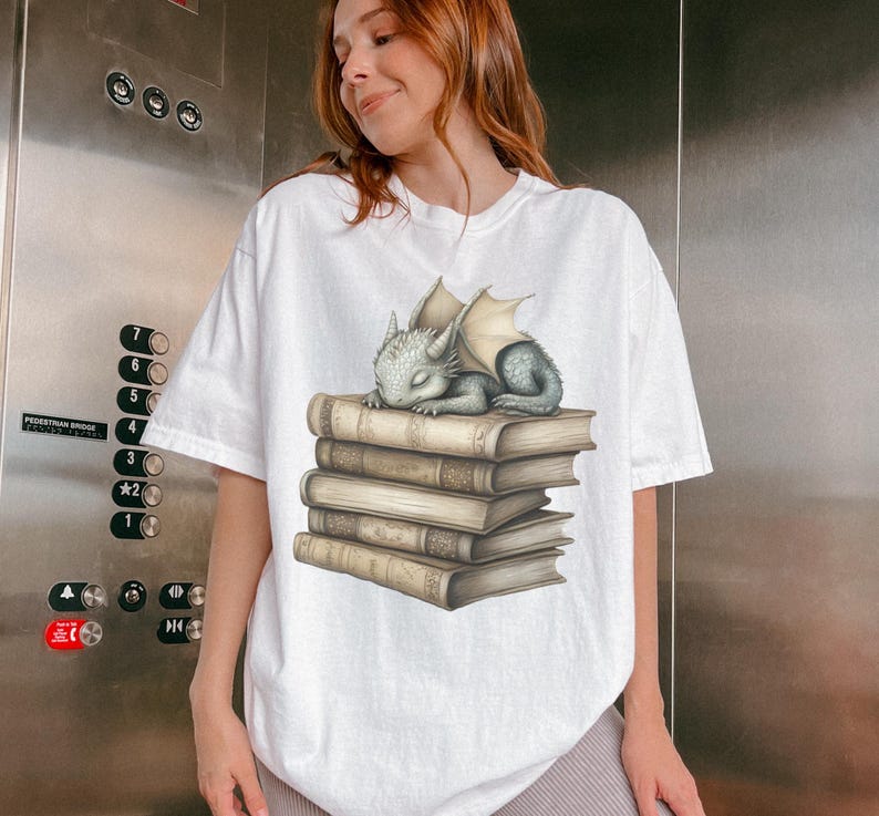 May include: White t-shirt featuring a sleeping baby dragon on a stack of books. The dragon is light blue with beige wings. The books are beige with detailed shading. The t-shirt is a loose fit.