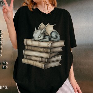 May include: Black t-shirt featuring a sleeping baby dragon illustration atop a stack of books. The dragon is gray with folded wings. The books are beige with detailed textures. The word "BLACK" is printed at the bottom.