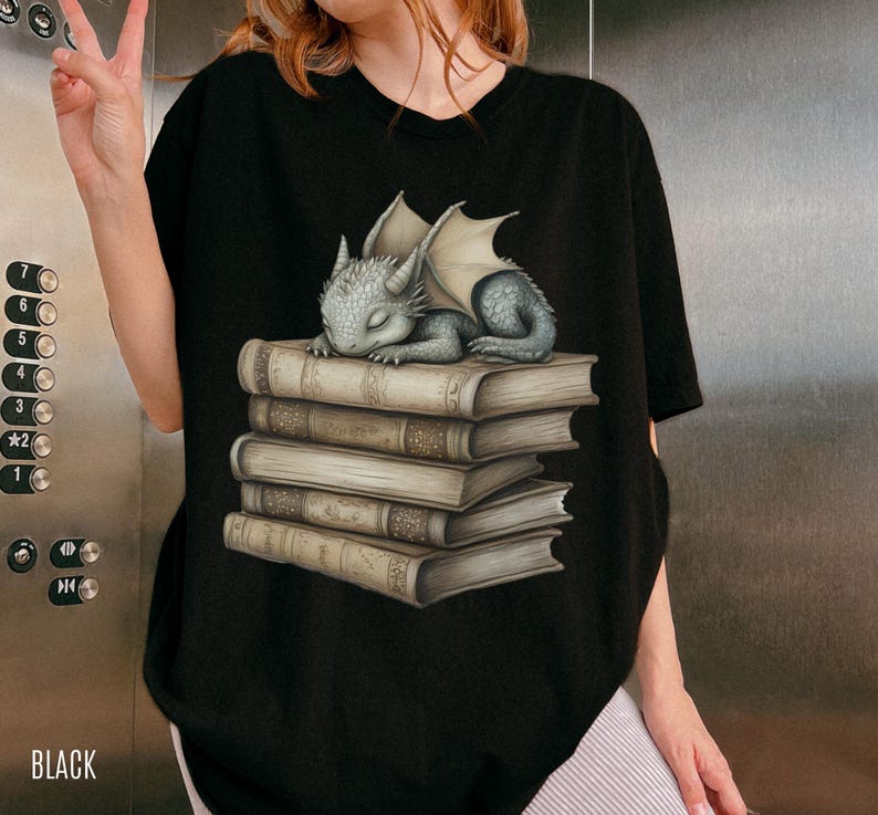 May include: Black t-shirt featuring a sleeping baby dragon illustration atop a stack of books. The dragon is gray with folded wings. The books are beige with detailed textures. The word "BLACK" is printed at the bottom.