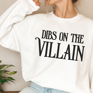K&ouml;nnte beinhalten: Wei&szlig;es Sweatshirt mit dem schwarzen Aufdruck "DIBS ON THE VILLAIN". Der Text ist fett und zentriert. Das Sweatshirt hat einen Rundhalsausschnitt und lange &Auml;rmel. Das Material scheint weich zu sein.