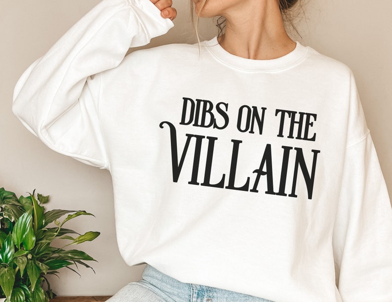 K&ouml;nnte beinhalten: Wei&szlig;es Sweatshirt mit dem schwarzen Aufdruck "DIBS ON THE VILLAIN". Der Text ist fett und zentriert. Das Sweatshirt hat einen Rundhalsausschnitt und lange &Auml;rmel. Das Material scheint weich zu sein.