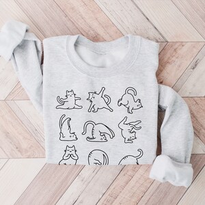 May include: A pale grey sweatshirt with a black outline illustration of cats performing yoga. The sweatshirt has a round neck and long sleeves with turned-up cuffs. The design is in the centre of the garment.