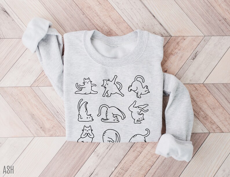 May include: A pale grey sweatshirt with a black outline illustration of cats performing yoga. The sweatshirt has a round neck and long sleeves with turned-up cuffs. The design is in the centre of the garment.