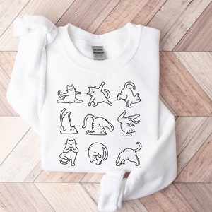May include: White sweatshirt with a black line drawing of cats in yoga poses. The cats are depicted in various positions, such as downward dog and lotus. The sweatshirt is made of a soft material and is suitable for casual wear.