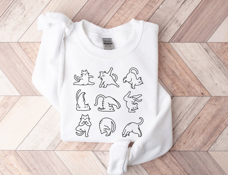 May include: White sweatshirt with a black line drawing of cats in yoga poses. The cats are depicted in various positions, such as downward dog and lotus. The sweatshirt is made of a soft material and is suitable for casual wear.