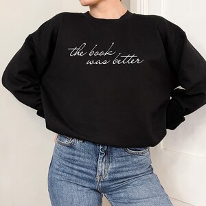 Puede incluir: Una sudadera negra de manga larga con cuello redondo que presenta el texto en cursiva blanca "the book was better" impreso en el pecho. La sudadera se combina con pantalones vaqueros azules, mostrando un estilo informal con cuello redondo y pu&ntilde;os acanalados.