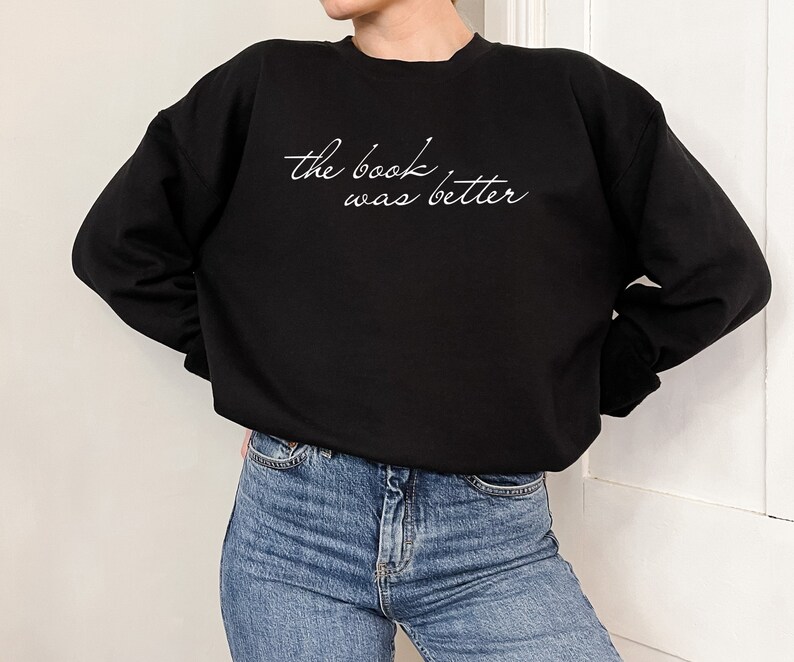 Puede incluir: Una sudadera negra de manga larga con cuello redondo que presenta el texto en cursiva blanca "the book was better" impreso en el pecho. La sudadera se combina con pantalones vaqueros azules, mostrando un estilo informal con cuello redondo y pu&ntilde;os acanalados.